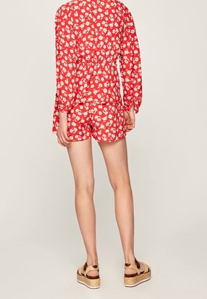 Kvinde iført en rød blomstret langærmet top og matchende shorts med beige platformsandaler, stående mod en ensartet lys baggrund.