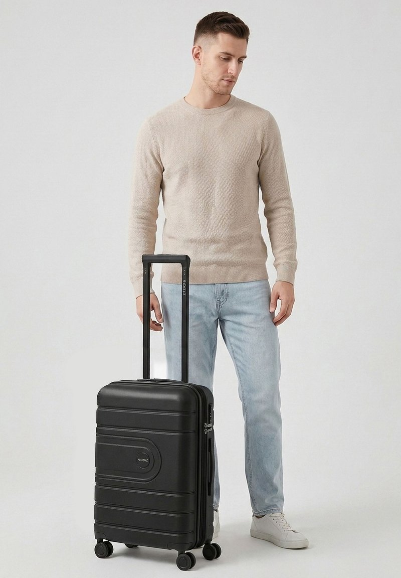 Giovane uomo con maglione beige e jeans chiari in piedi che tiene una valigia nera con ruote e manico esteso.