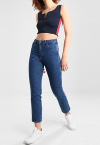 Top corto blu navy con zip e striscia rossa, abbinato a jeans blue high-waisted. Il modello indossa sneakers bianche; tessuto liscia e design aderente.