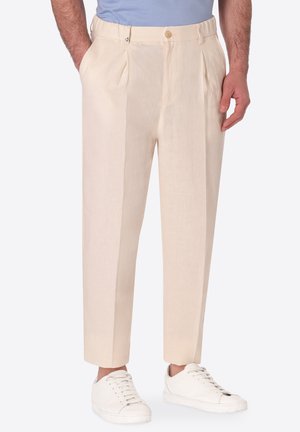 Uomo che indossa pantaloni beige plissettati con tasche laterali e sneakers bianche su uno sfondo chiaro e uniforme.