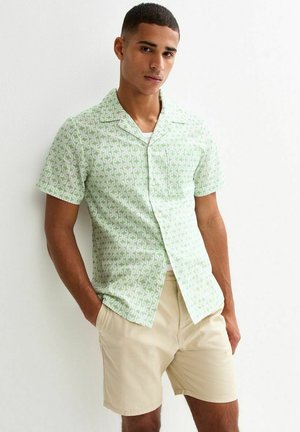 Camicia a maniche corte in tessuto verde chiaro con un motivo astratto ripetuto; abbinata a pantaloni beige neutri; texture liscia.