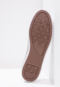 Sole bílé tenisky Converse All Star s hnědou gumovou podrážkou, umístěná vertikálně proti bílé rohové zdi.