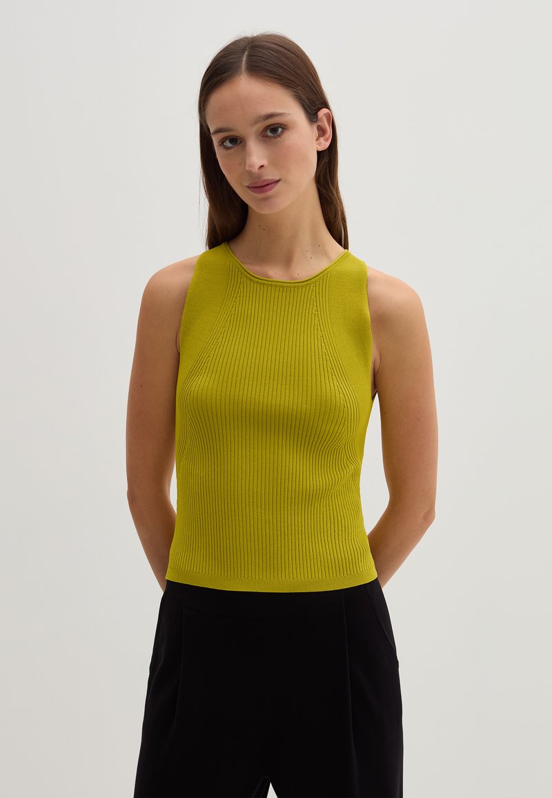 Giovane donna con lunghi capelli castani, che indossa un top senza maniche lime verde a coste e pantaloni neri, in piedi di fronte a uno sfondo chiaro e semplice.
