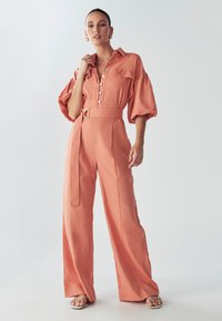 Mono de lino melocotón con cierre de botones en la parte delantera, mangas abullonadas, cintura ceñida con cinturón y piernas anchas. Notable bolsillo de parche en el pecho.