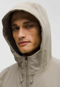 Chaqueta beige con capucha y cuello alto, que presenta un cierre frontal y detalles de costura sutiles. Textura suave y lisa, diseño minimalista.