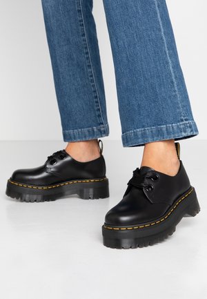 Personne portant des chaussures à plateforme épaisses noires avec des coutures jaunes et un jean bleu denim, debout sur un sol clair.
