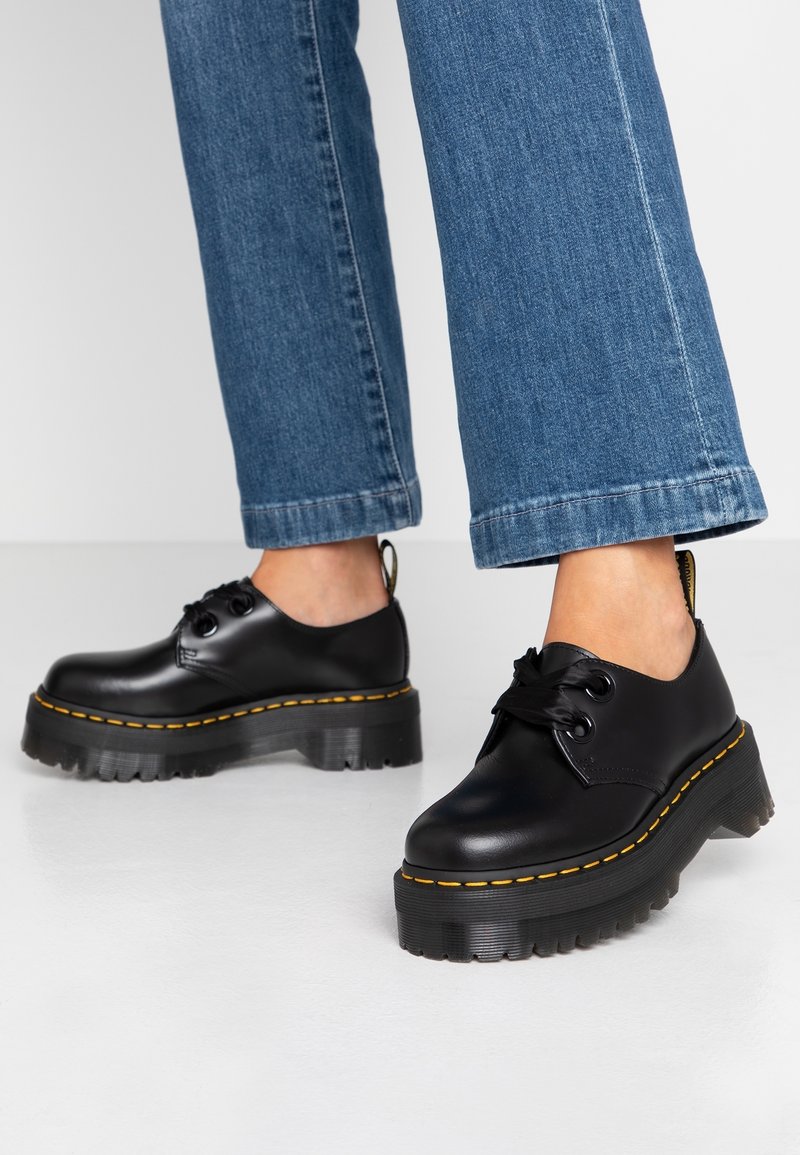 Personne portant des chaussures à plateforme épaisses noires avec des coutures jaunes et un jean bleu denim, debout sur un sol clair.
