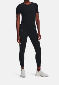 Osoba ubrana w czarną, krótką sportową koszulkę i legginsy z małymi logo oraz białe sneakersy, stojąca na jednolitym białym tle.