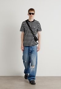 Black and white striped t-shirt, relaxed fit, paired with light blue distressed jeans and black shoes; model wears sunglasses and a crossbody bag.