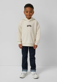 Crème hoodie met gestikte "SLVR" logo, donkerblauwe jeans en witte sneakers met grijze accenten. Glad materiaal en ontspannen pasvorm.