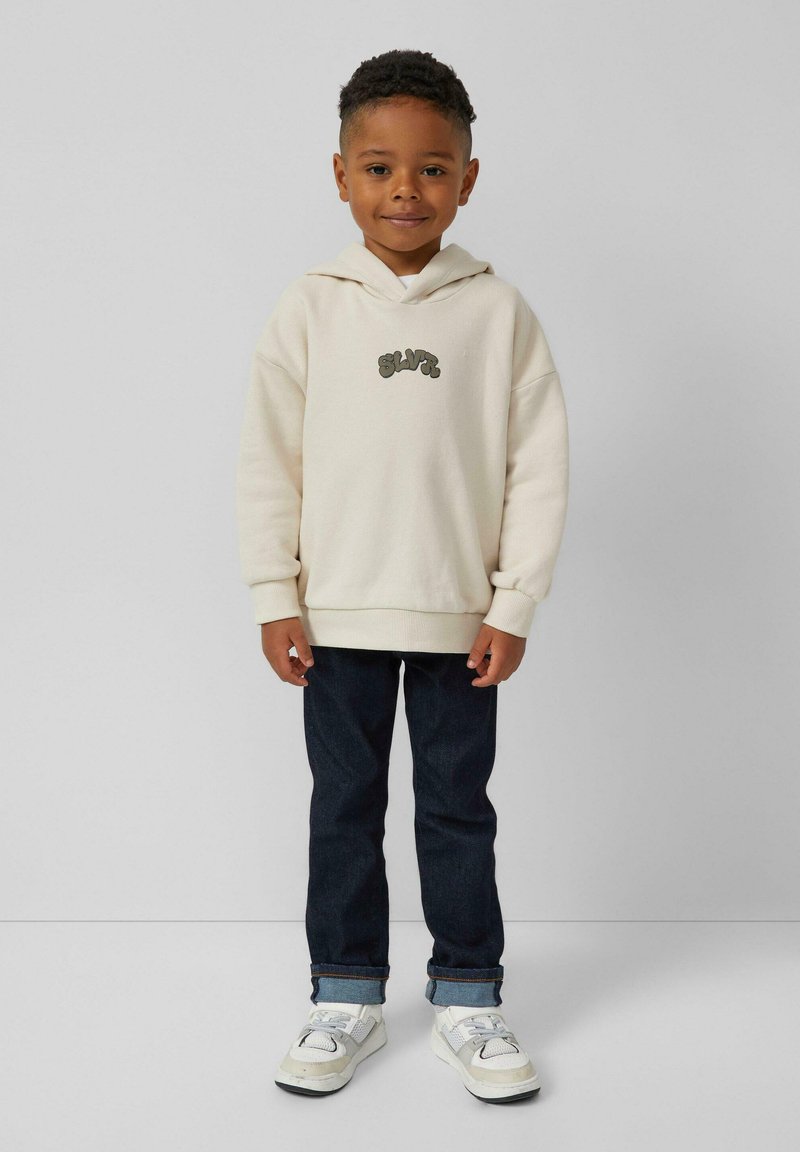 Crème hoodie met gestikte "SLVR" logo, donkerblauwe jeans en witte sneakers met grijze accenten. Glad materiaal en ontspannen pasvorm.