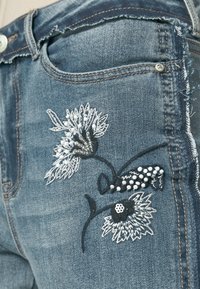 Siniset farkut, joissa on valkoista ja mustaa kukkaistikkausta, koristeellisia helmikoristeita ja nyppyinen reunusdetaili.