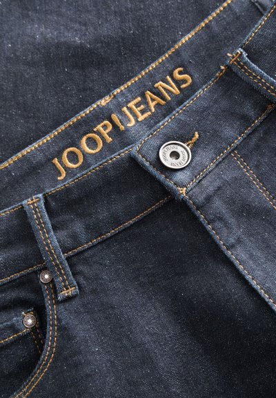 Jean en denim foncé avec des coutures jaunes, featuring un bouton en métal et "JOOP JEANS" brodé au-dessus de la ceinture. Texture lisse.