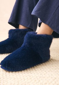 Scarpette morbide blu navy realizzate in materiale soffice con un gambale alto. Texture robusta sulla parte esterna e suola interna liscia. Posate su un tappeto chiaro.