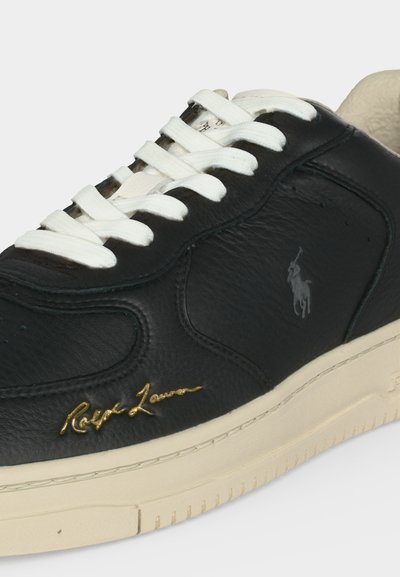 Μαύρο δερμάτινο sneaker με λευκά κορδόνια, κρεμ σόλα, χρυσή υπογραφή "Ralph Lauren" και γκρι λογότυπο παίκτη πόλο στο πλάι.