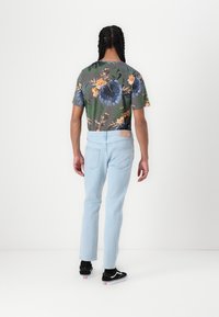 Menns avslappede lyseblå jeans kombinert med en kortermet skjorte med mørk blomstermønster. Svart sneakers med hvite såler. Bakfra.