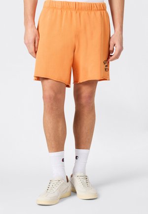 Hombre con pantalones cortos naranjas con cintura elástica y texto negro en la pierna izquierda, calcetas blancas hasta la mitad de la pantorrilla y zapatillas off-white sobre fondo blanco.