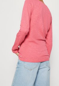 Langärmliges Oberteil aus melange-pinkem Stoff mit einem Bootsausschnitt, charakterisiert durch eine lockere Passform und ein dezentes Markenlogo nahe dem Saum. Kombiniert mit hellen Jeans.