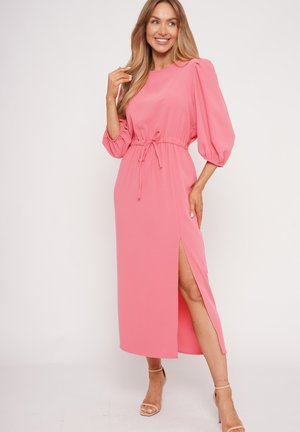 Woman wearing a long pink dress with puff sleeves, waist tie, and side slit, standing in neutral heels, smiling against a plain background.