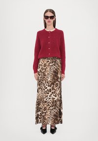 Cardigan rosso a maglia con dettagli a bottone, abbinato a una lunga gonna con stampa leopardata. Il modello indossa occhiali da sole e décolleté neri a punta.