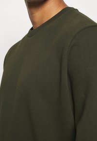 Tmavě zelená mikina s kulatým výstřihem, měkkou texturou a žebrovaným límcem. Detailní pohled zdůrazňující látku a design výstřihu.