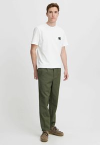 T-shirt en coton blanc avec une petite poche logo noire, associée à un pantalon vert olive. Des chaussures à lacets marron complètent l'ensemble.
