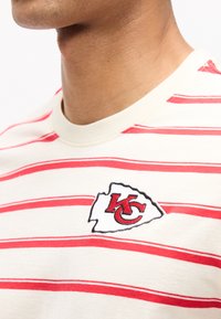 Posnetek od blizu osebe, ki nosi belo-rdečo črtasto majico z logotipom Kansas City Chiefs na prsih.