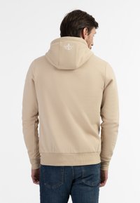 Beige hættetrøje lavet af blødt stof, med snoretræk i hætten, ribbede manchetter og et logoaccent på ryggen. Parret med blå jeans.