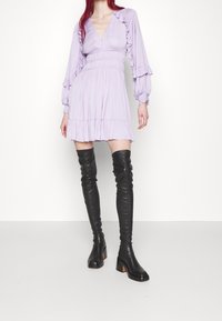 Lila mini-jurkje met ruches aan de mouwen en een V-hals, gecombineerd met zwarte over-de-knie leren laarzen met dikke hakken.