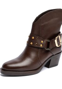 Bota de cuero marrón hasta el tobillo con punta afilada, tacón apilado y una correa decorativa con tachuelas doradas y hebilla. Textura lisa.