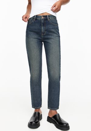 Personne portant un jean bleu délavé droit, une chemise blanche légèrement relevée et des chaussures noires slip-on à plateforme épaisse.
