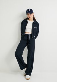 Granatowy dres składający się z bluzy zapinanej na zamek i spodni o szerokich nogawkach z białym sznurkiem, zestawiony z niebieską czapką z logo H i białymi trampkami.