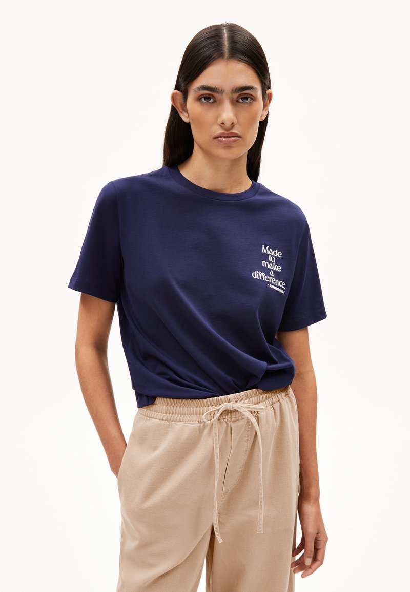 Frau mit langen, dunklen Haaren trägt ein navyblaues T-Shirt mit dem Text "Made to make a difference" und beigen Jogginghosen mit Kordelzug, steht vor einem weißen Hintergrund.