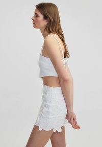 Top corto blanco con tirantes finos y espalda descubierta, combinado con una falda mini blanca que presenta bordados florales y un dobladillo en forma de ondas.