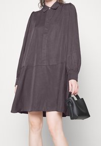 Abito button-up a maniche lunghe di colore grigio scuro con una silhouette rilassata e spalle arricciate. Abbinato a una mini borsa nera.