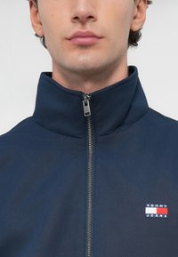 Giacca a zip blu navy con colletto alto, caratterizzata da hardware in argento e un piccolo logo Tommy Jeans multicolore sul lato sinistro del petto.