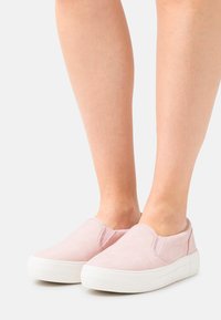 Sapatos de slip-on cor-de-rosa com uma textura de camurça suave, sola de borracha branca e detalhes elásticos laterais projetados para fácil calçar.