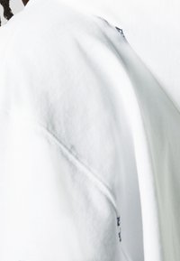 Witte trui met een gladde textuur, voorzien van een ronde hals en doorgestikte naden, met een subtiel grafisch detail vlak bij de schouder.