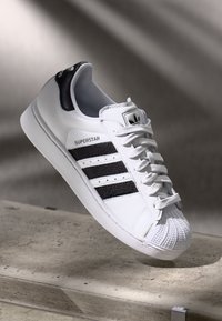 Biele tenisky Adidas Superstar s čiernymi pruhmi a špičkou v tvare mušle odložené na betónovom okraji s rozmazaným pozadím.