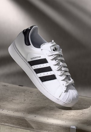 Sneaker Adidas Superstar bianco con strisce nere e punta a conchiglia appoggiata su un bordo in cemento contro uno sfondo sfocato.