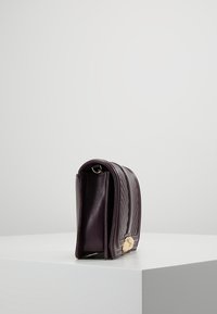 Borsa a tracolla in pelle viola con design trapuntato, caratterizzata da una superficie liscia e un accento in metallo dorato sul lato. Vista laterale in evidenza.