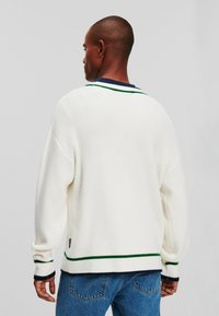 Suéter blanco de punto con textura acanalada, con rayas verdes y azul marino en el cuello y el dobladillo. Presenta un ajuste holgado y mangas con caída.