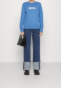 Person som bär en blå sweatshirt med texten "BOSS", uppvikta blå jeans, svarta plattformsskor och håller en svart handväska med runda handtag.