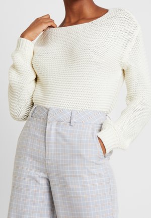 Personne portant un pull en maille épaisse blanc et un pantalon à carreaux bleu clair taille haute, une main dans la poche et l'autre touchant le col.