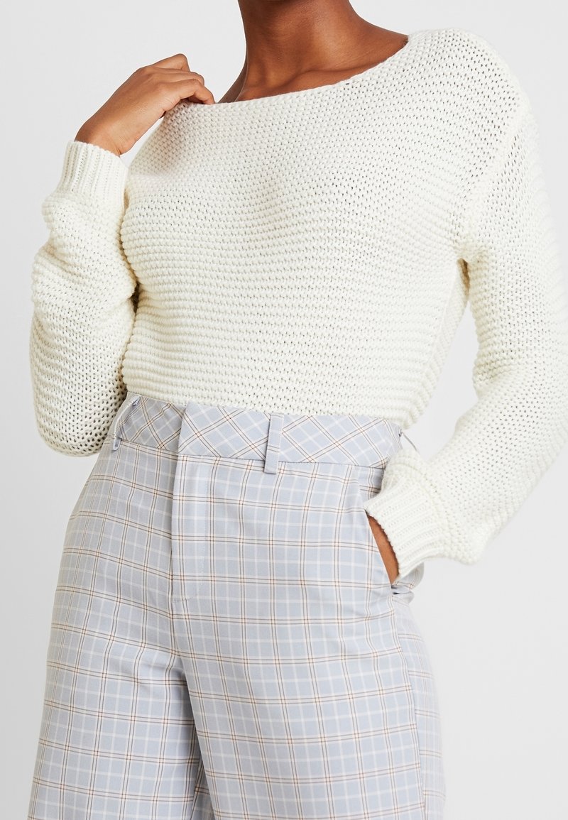 Personne portant un pull en maille épaisse blanc et un pantalon à carreaux bleu clair taille haute, une main dans la poche et l'autre touchant le col.
