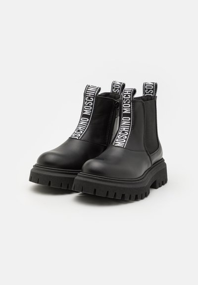 Botas de tobillo de cuero negro con paneles laterales elásticos, suela de goma gruesa y tiradores con marca que presentan "MOSCHINO" en blanco sobre un fondo negro.