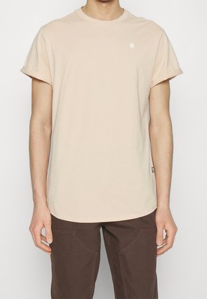 Homme portant un t-shirt à manches courtes beige clair uni avec un petit logo sur la poitrine, associé à un pantalon marron foncé, sur fond blanc.