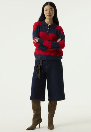 Mujer con el cabello largo y oscuro lleva un jersey de rayas rojas y azul marino, pantalones cortos de mezclilla de pierna ancha, botas altas marrones y un pañuelo negro atado a los shorts.