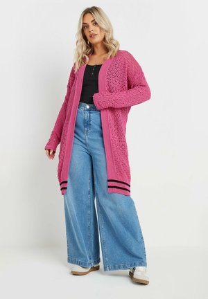 Cardigan tricoté rose avec un design texturé, manches longues, devant ouvert et poignets rayés ; associé à un jean bleu à large jambe et des baskets blanches.
