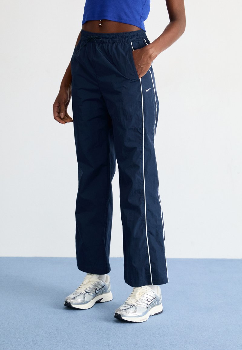 Marineblauwe windjackbroek met witte zijstrepen, elastische tailleband en zijzakken, gemaakt van lichtgewicht, glad materiaal.